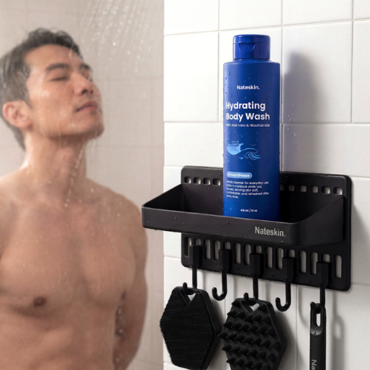 A man is shown under a shower with water running, while a blue bottle of Nateskin Hydrating Body Wash is displayed on a black wall-mounted shower shelf with hanging brushes.